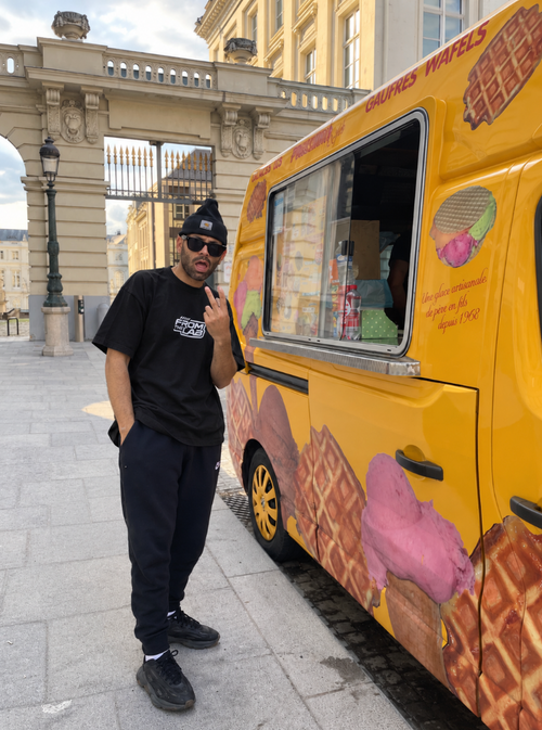Alexander T. Rose wearing The black  From The Lab t-shirt,stands next to a bright yellow food truck decorated with waffles and ice cream graphics, set against a historic stone building background. in Brussels Belgium
